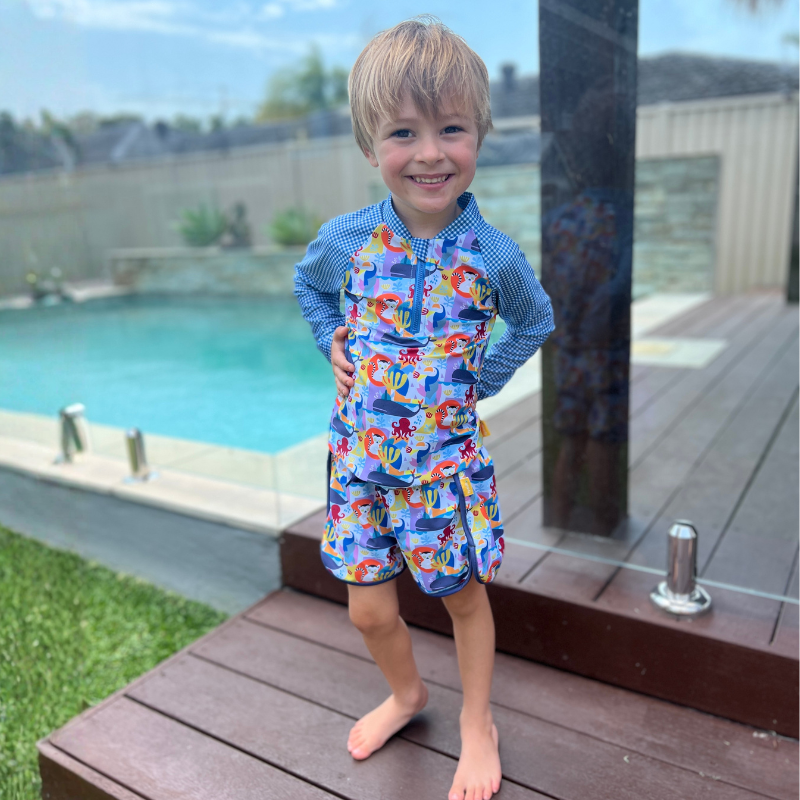 young boy age 5 stands in tyoub animal cutout print swim shorts and tyoub animal cut out long sleeve rashguard next to a swimming pool.