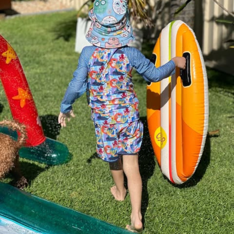 Child in a tyoub animal cutout print swim shorts and rash top swim set and tyoub skulls sunhat holding an inflatable board on grass. 